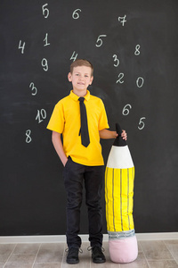 可爱的英俊的学校男孩在黄色 tshirt 领带并且时髦的靴子休闲站立 cloase 到黑板与数字和拿着巨大的大软的玩具铅笔和微
