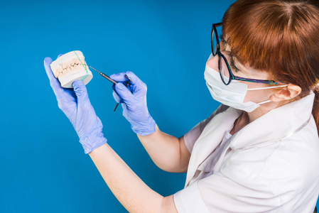 成人经验丰富的红头发的妇女医生牙医在白色的保护性医疗口罩的脸和眼镜持有一个石膏铸造的牙齿在手