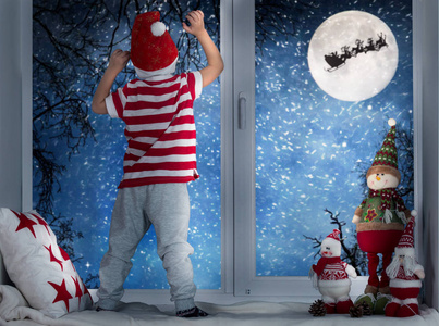 圣诞快乐, 节日愉快小男孩站在窗前, 看着圣诞老人在雪橇上飞向月亮的天空。在圣诞节装饰的房间。孩子享受假期
