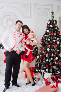 女婴穿着可爱的红色连衣裙和圣诞老人与母亲在红礼服和父亲, 附近的圣诞树在载歌载舞装饰房间与花环的灯光。圣诞节和新叶的白色和红色的