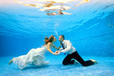 一个男人和一个女孩穿着婚纱拥抱在水下，坐在游泳池的底部。 水平视图。 水下射击