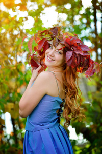 阳光明媚的一天，一个美丽的女孩，留着一头长发，穿着蓝色的连衣裙，戴着一个红色和绿色的叶子花环，站在明亮的秋天灌木丛中，侧过肩膀，