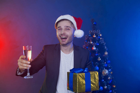 新年好，圣诞快乐。 美丽微笑的年轻商人戴着圣诞老人的帽子，手里拿着一杯香槟和一份礼物，背景是圣诞树。