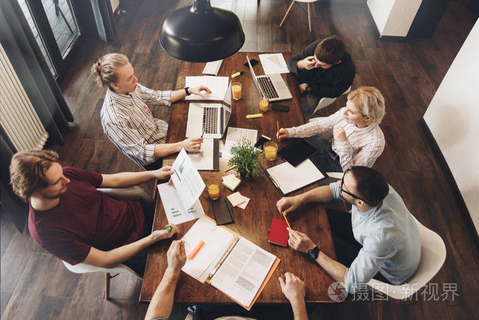 一组创造性的同事围坐在一个开放的空间的木桌旁, 与商业文档和笔记本