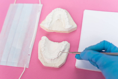 在粉红色的背景是一个白色的保护面具, 石膏铸造牙齿, 牙医医生持有牙科仪器和工作