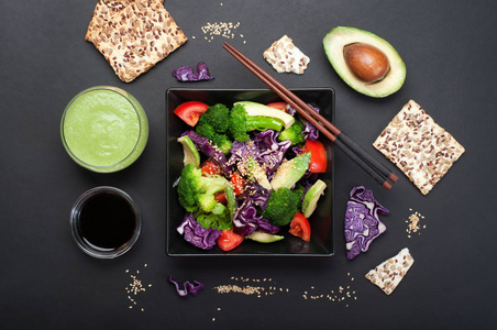 沙拉从花椰菜, 紫罗兰菜, 西红柿和鳄梨与蜂蜜酱和橄榄油在一个黑色的方形板上的黑暗背景。靠近绿色的果汁和酱油。健康膳食营养的概念