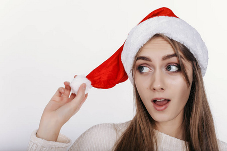 戴着圣诞帽的女人侧身看着令人兴奋的。 漂亮的年轻模特，金色的长发，宽眉，蓝色的眼睛在白色背景的演播室里摆着相机。 孤立的。 复制