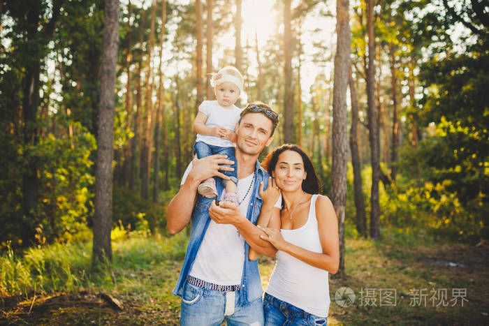 时尚的年轻家庭的妈妈, 爸爸和女儿一岁的金发碧眼坐在靠近父亲的肩膀, 户外在城市外的公园里, 在夏天的大树在日落
