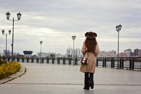 有吸引力的时髦女孩穿着长大衣带和大帽子漫步在长廊上。秋天在清新的空气中漫步