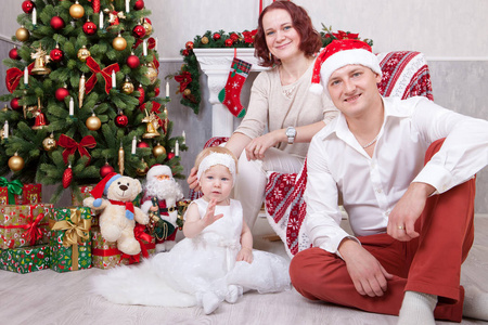圣诞节或新年庆祝。三人的快乐年轻家庭的画像在圣诞节圣诞树附近与圣诞节礼物。背景上有圣诞袜的壁炉。节日快乐