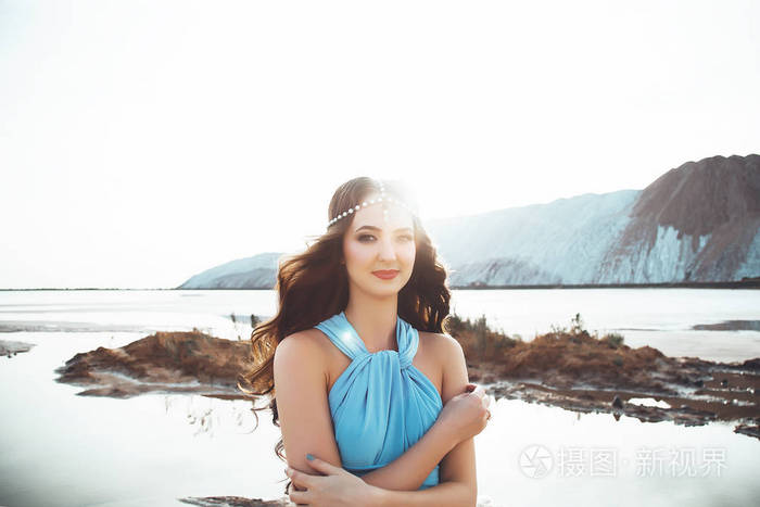 迷人的白种女人的肖像与长的黑发, 俏丽的女孩在蓝色礼服和与王冠在头发反对风景的美丽的背景, 妇女在湖附近和
