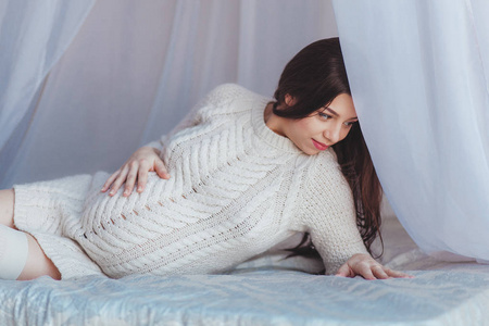 孕妇针织美丽的怀孕妇女躺在床上的树冠.穿白色针织套衫.产科概念照片
