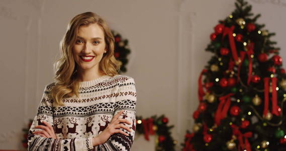 年轻的金发美女的肖像拍摄真诚的微笑, 并与交叉的手看着相机。带有红色丝带和灯光背景的圣诞树。室内.关闭