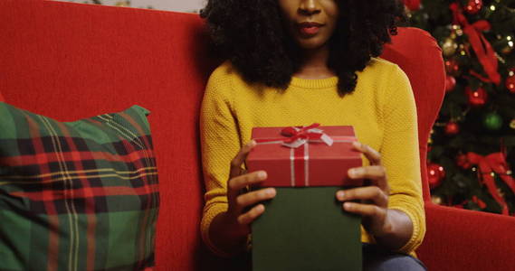 非洲裔美国青年卷曲的妇女打开圣诞节礼物与红色丝带在它和有惊奇愉快的面孔, 当坐在红色长沙发在客厅里。圣诞树灯