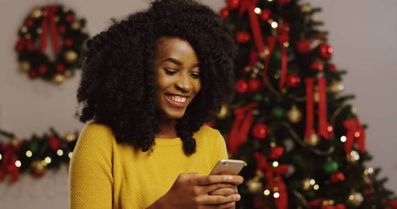 结束了年轻的美国黑人卷曲的女人在她的智能手机上录音和笑。圣诞树上有灯光的背景。室内.肖像拍摄