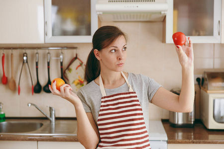一个年轻的困惑和沉思的女人在围裙决定选择一个红色或黄色的西红柿在厨房。节食概念。健康的生活方式。在家做饭。准备食物