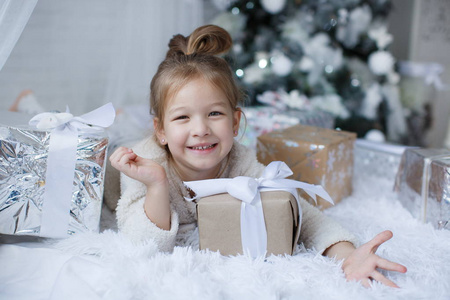 快乐的孩子5岁的女孩在圣诞节假期靠近一棵美丽的绿色圣诞树，上面装饰着白色的玩具和雪花，旁边放着白色地板上的大盒子里漂亮的圣诞礼物