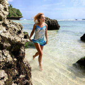 带着丝质衣服的快乐金发女郎微笑着走在巴厘岛的狂野海岸上