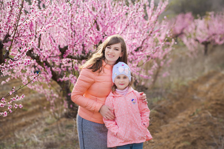 可爱的漂亮时髦的黑发女孩, 妈妈站在春天的田野上, 一棵粉红色的小花。穿雨衣的女士和蓝色帽子橡胶靴子春天季节