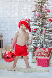 英俊的男孩在红色圣诞老人温暖的帽子与大红色圣诞树玩具球庆祝新年接近圣诞节树充满礼物