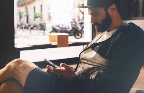 一个留胡子的嬉皮士正在寻找通过他的智能手机上的社交网络应用程序与他们的朋友聊天。一张年轻英俊的自由职业者拿着手机坐在桌子旁的照片