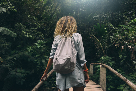 从照片的背面看，年轻的旅游女性卷曲的头发走在老木桥在丛林中。背包族女孩正在暑假探索热带森林..