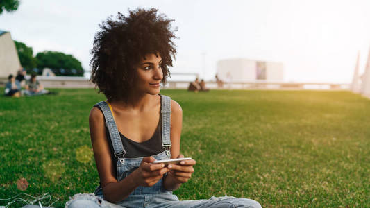 有非洲发型的旅行博主女性坐在户外，手里拿着手机，微笑着。 年轻的企业家正在通过连接wifi的智能手机与合作伙伴在线聊天。