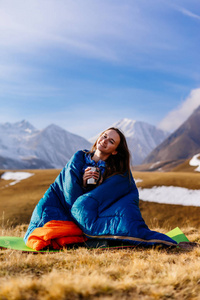 一个微笑的年轻女孩拿着一个杯子与热茶, 坐在一个睡袋里反对高加索山的背景, 喜欢旅行
