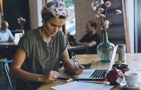 年轻时尚的女人在现代咖啡店工作时，正在用手机听音乐。 企业家正在进行一个新的商业项目，一台笔记本电脑连接到wifi。