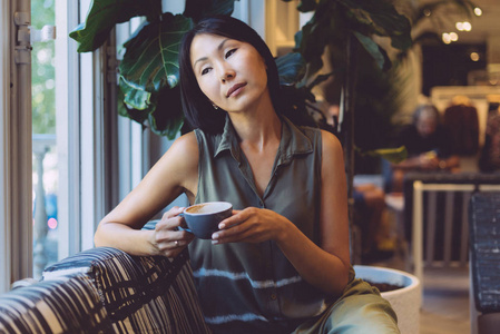 美丽的亚洲女孩，留着长长的黑发，坐在舒适的咖啡馆里，享受着早晨的咖啡。 一位年轻女性穿着休闲服，坐在咖啡店里喝杯咖啡。
