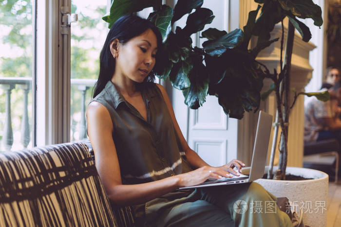 年轻的亚洲女人坐在现代咖啡店里，用笔记本电脑打字电子邮件。 自由职业者女孩正在与客户在线沟通，同时访问现代合作空间。