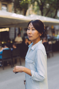 年轻的模特看起来是亚洲女性，穿着牛仔裤，站在一边，站在模糊的街道背景上。 现代博主正在周末短途旅行中探索城市的新景点。