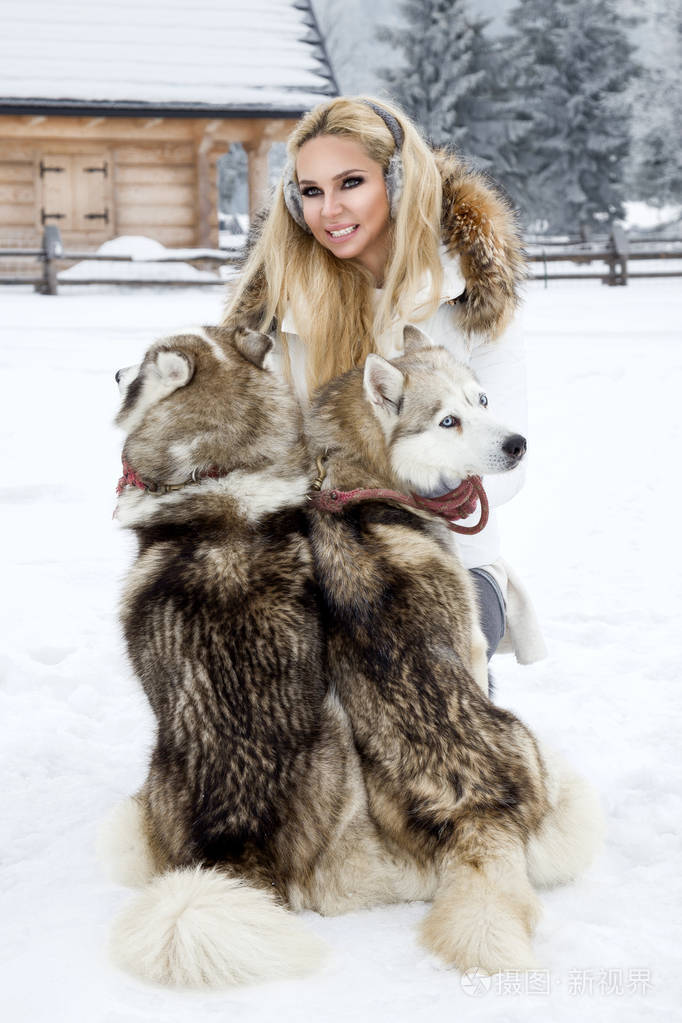 美丽的金发女人站在雪地上抱着哈士奇的狗背景是美丽的山和雪的景色