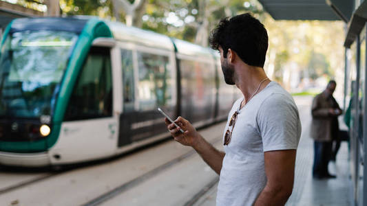 胡须男，黑发短信在手机上，而站在城市汽车站。 时髦的家伙穿着休闲服装，在连接wifi的智能手机上上网。