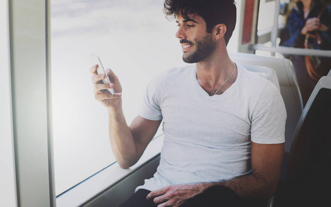 留胡子的家伙在一台智能手机的显示器上阅读电子邮件，它连接到公共Wi Fi，而坐在城市公共汽车上。 年轻的帅哥在发短信的时候看着手