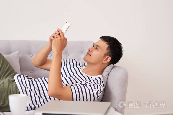 轻松的亚洲男人用智能手机躺在家里客厅的沙发上