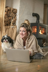 客厅里年轻漂亮的女人坐在地板上，通过电脑和朋友交流，用毯子盖着壁炉后面的寒冷