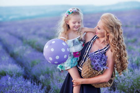 年轻的母亲带着年轻的女儿在薰衣草的田野上微笑。女儿坐在母亲的手上。穿彩色连衣裙的女孩, 穿着深蓝色连衣裙的母亲