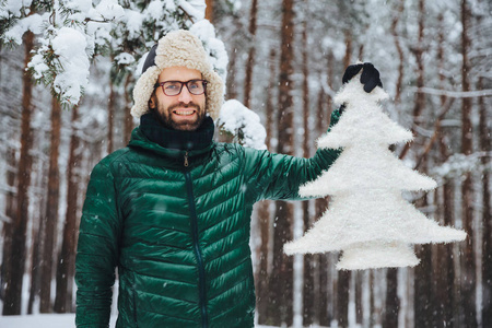 穿着暖和衣服的英俊的长胡子的男人, 捧着人工枞树, 站在白雪覆盖的美丽的树上, 有欢快的积极表达, 呼吸着清新的冬日空气