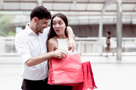 在亚洲国家进行了一天的大减价购物之后，快乐的一对夫妇在交谈。年轻英俊的白人男性和美丽的亚洲女性带着红色的购物袋，看上去非常满意。