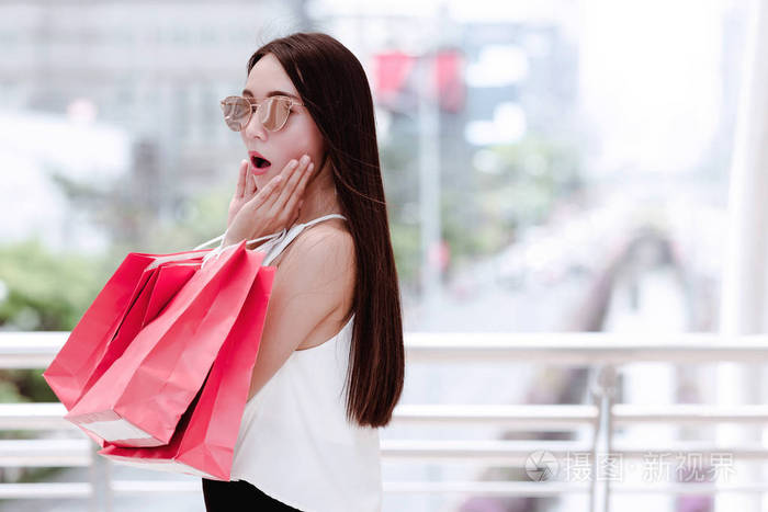 年轻的购物狂女人的肖像，有许多红色购物袋，有惊喜的脸，因为她看到一个销售标志。 在自然光下室外拍摄。