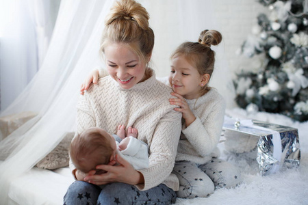 快乐的女人，快乐的母亲和小孩子，一个女孩和一个新生的男孩，坐在圣诞喜庆的绿色圣诞树旁边，白色玩具，雪花白色的蝴蝶结和发光的灯。
