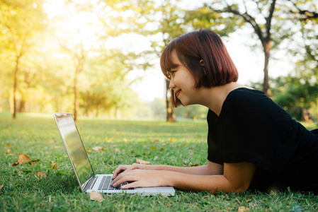 年轻的亚洲女子的腿在绿色的草地上, 打开笔记本电脑。女孩的手在键盘上。远程学习概念。在公园里工作的笔记本电脑的快乐的时髦的年轻亚