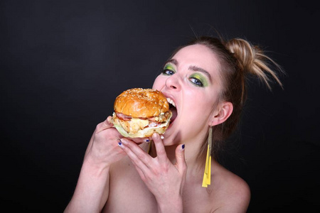 那个女人正在吃一个带肉的巨型汉堡包。 在家里送食物的胃口午餐。 明亮的绿色时尚构成眼影黄色耳环。 横幅概念不健康的食物吃。