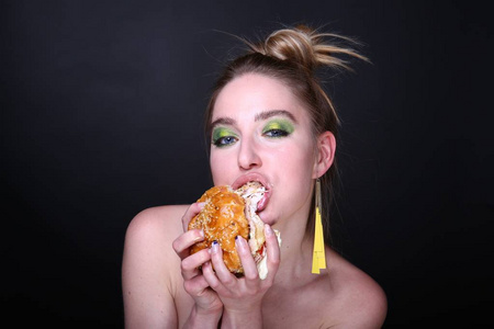 那个女人正在吃一个带肉的巨型汉堡包。 在家里送食物的胃口午餐。 明亮的绿色时尚构成眼影黄色耳环。 横幅概念不健康的食物吃。