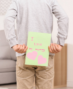 逗人喜爱的小男孩掩藏问候卡片为母亲节在他的后面在家