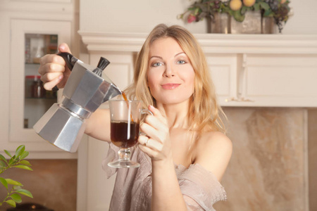 美丽的白种人金发家庭主妇站在家里的厨房里，拿着水壶和一杯早咖啡