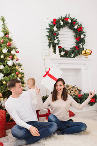 快乐的年轻开朗的父母带着可爱的小儿子。小男孩坐在家里的光房里, 带着装饰一新的年树和礼品盒在头上。圣诞节好心情。家庭, 爱和假日