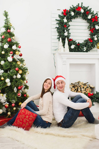 快乐欢快的滑稽的年轻情侣在红帽子坐在家里与装饰的新年树和礼品盒的背景下, 圣诞花环的光房间。家庭, 假日2018概念