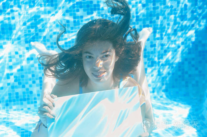 美丽的年轻女孩在游泳池里拿着白色的白板，在水中健身，享受家庭度假的乐趣。